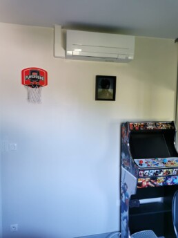 Unité intérieure murale Mitsubishi Electric MSZ-AP blanche, installée dans une salle de jeux au-dessus d’un mini panier et d’une borne d’arcade.